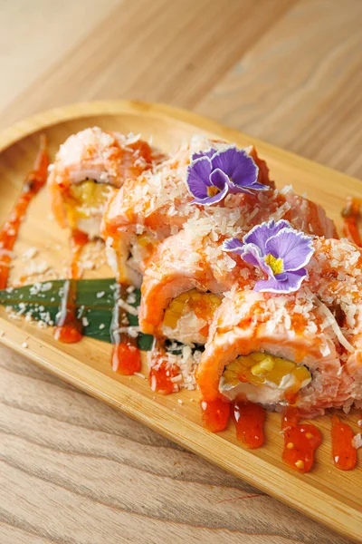 Renkli suşi ruloları ahşap bir tabakta güzelce düzenlenir. Tabakta, yenilebilir çiçeklerle süslenmiş taze malzemeler ve tat vermesi için soslar yer alıyor..