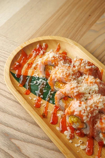 Lokantalar baharatlı sos ve çıtır çıtır parçalarla kaplı bir tabak suşi rulosunun keyfini çıkarıyorlar. Sunumda, tabağın cazibesini vurgulayan yeşil yapraklı ahşap bir tabak yer alıyor..