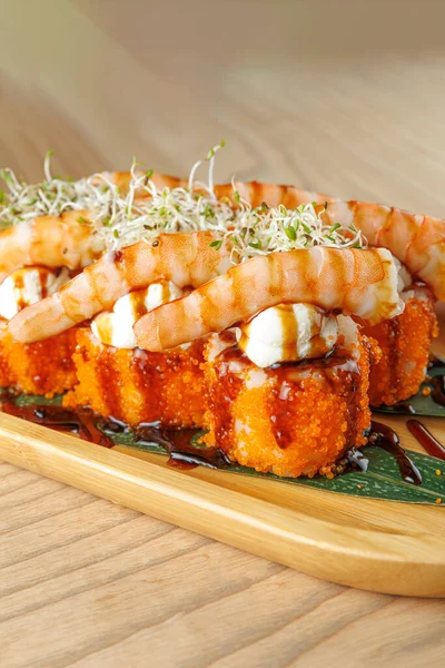 Bir tabakta karides ve sosla kaplı renkli suşi ruloları var. Tabak canlı renkler ve dokular sergiliyor. Seyirciyi Japon mutfağının tadına bakmaya çağırıyor..