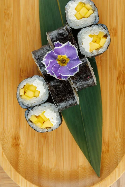 Taze mango parçalarıyla dolu suşi ruloları ahşap bir tabakta düzenlenir. Mor bir çiçek bambu yaprağının üzerine dekoratif bir dokunuş ekler. Bu yemek lezzet karışımı sunar..