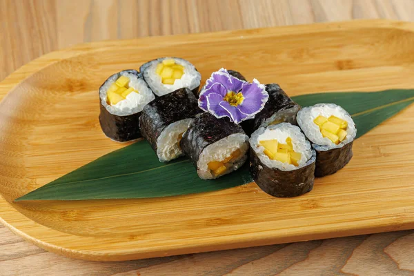 Suşi ruloları, altında yeşil bir yaprak olan bambu bir tabakta düzgünce düzenlenir. Ekmekler mango içeriyor ve süslemek için bir çiçekle kaplanmış..
