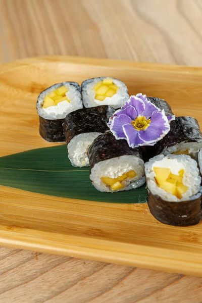 Krem peynirli lezzetli suşi ruloları ve ahşap bir tabağa yerleştirilmiş mango parçaları. Narin bir mor çiçek yemeğe biraz renk katar..