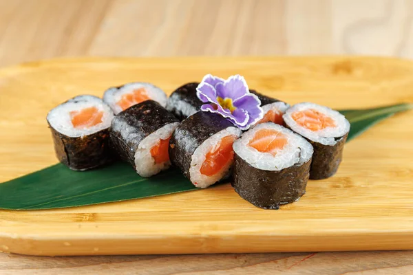 Somon ve pirinçle dolu suşi ruloları ahşap bir tahtada güzelce düzenlenir. Yenilebilir bir çiçek sunuma biraz renk katar ve çekici bir yemek yaratır..
