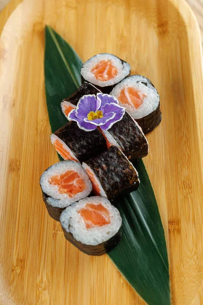 Bir bambu tabağında taze somonla dolu çeşitli suşi ruloları düzenlenir. Mor bir çiçek sunuma renkli bir dokunuş ekler. Bu görüntü Japon mutfak sanatını ön plana çıkarıyor.