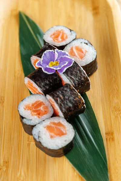 Somon balıklı suşi ruloları bambu tabakta güzelce düzenlenir. Mor yenilebilir bir çiçek yemeğe renk katar. Dekor, taze malzeme ve işçiliği ön plana çıkarıyor..