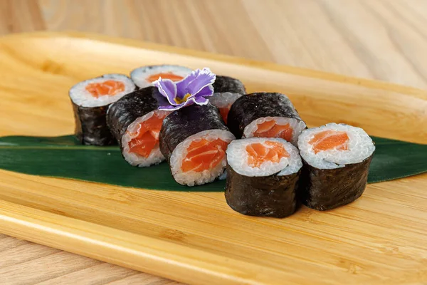 Suşi ruloları ahşap servis tabağına düzgünce yerleştirilir ve deniz yosununa sarılmış taze somon parçaları sergilenir. Küçük bir çiçek sunuma biraz stil katar.