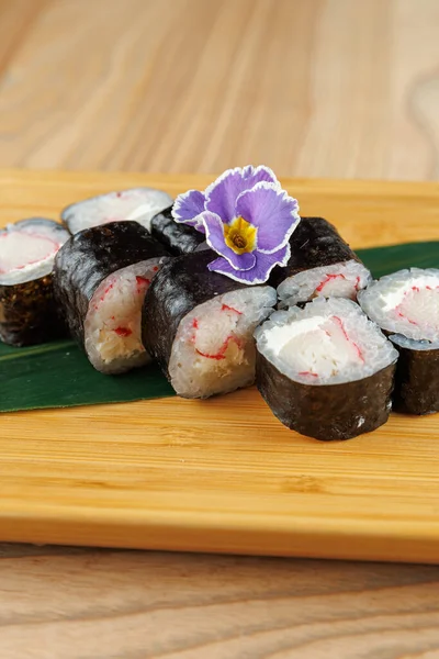 Taze suşi ruloları ahşap bir tahtada renkli bir çiçek süslemesi ile düzenlenir. Ekmekte deniz yosunu, pirinç ve dolgu var. Canlı bir yemek seçeneği yaratıyor..