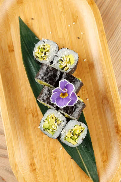 Deniz yosunu, pirinç ve sebzelerle yapılan suşi ruloları bambu bir tabakta sergilenir. Mor bir çiçek ve yeşil bir yaprak ayara renk katar.