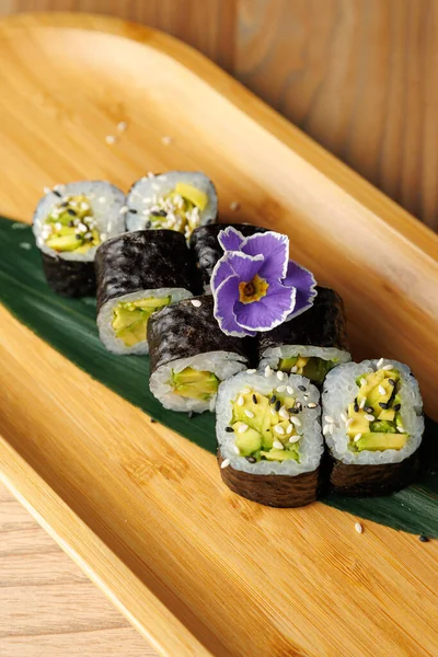 Salatalık ve avokado ile dolu suşi ruloları yeşil bir yaprak üzerinde güzelce düzenlenir. Bir çiçek, ahşap tabağın sunumuna renk ve tazelik katar..