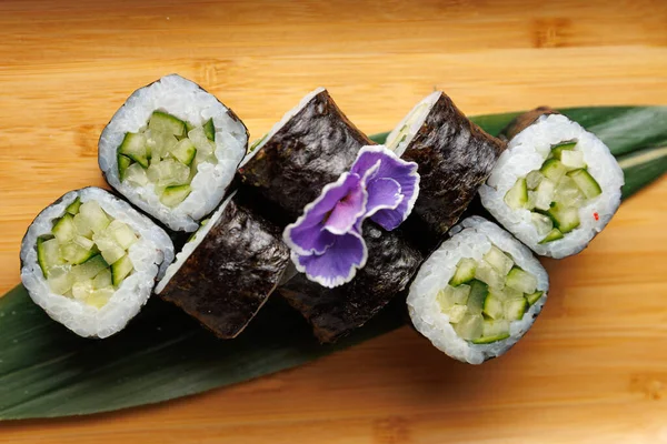 Taze salatalıkla dolu suşi ruloları bambu yüzeyine yerleştirilmiş bir yaprağa yerleştirilmiştir. Mor bir çiçek sunuma biraz renk katar.