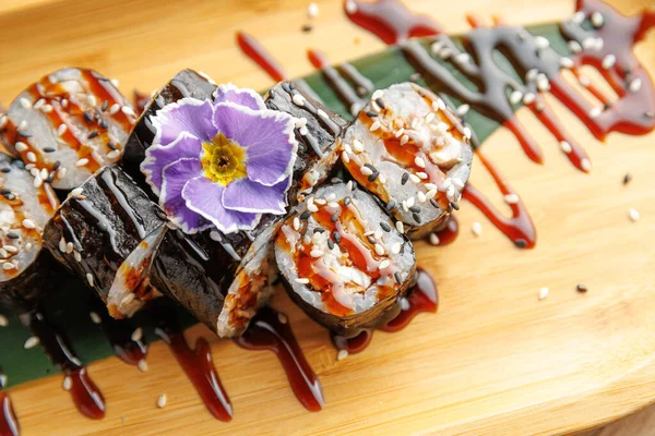 Ahşap bir tabakta sergilenen suşi ruloları bir çiçek süslemesi ve lezzetli bir çiseleme içeriyor. Yemek Japon mutfağından tatlar ve görselleri birleştiriyor..