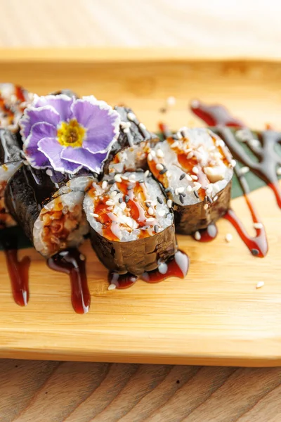 Suşi ruloları ahşap bir tabakta güzelce düzenlenir. Ekmekler susam tohumlarıyla kaplı ve sosla ıslatılmış. Mor bir çiçek tabağa dekoratif bir dokunuş ekler..