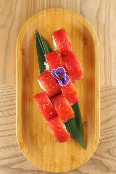 Suşi ruloları yeşil yapraklı bir bambu tabakta ince düzenlenir. Üzerinde küçük mor bir çiçek oturuyor, sunuma biraz renk katıyor..