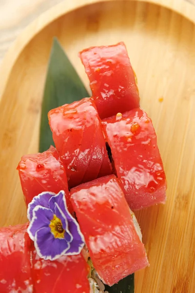 Taze ton balığıyla dolu suşi ruloları ahşap bir tabakta servis edilir. Yenilebilir bir çiçek sunuma renk katar. Bu yemek malzemelerin kalitesini ve tazeliğini vurgular..