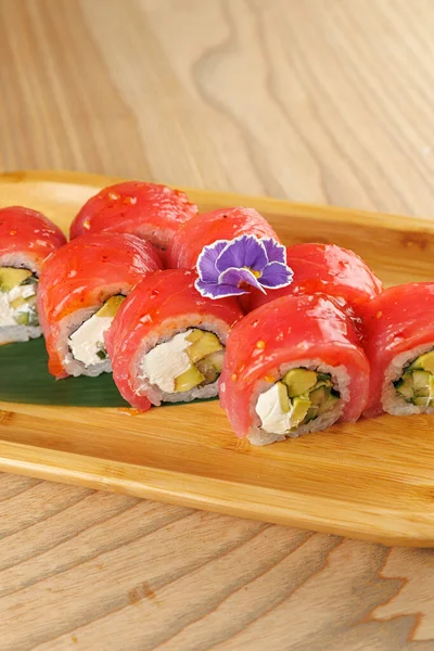 Avokado ve krem peynirle dolu suşi ruloları ahşap bir tabakta güzelce düzenlenir. Mor bir çiçek sunuma renk ve tazelik katar.