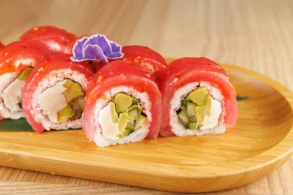 Suşi ruloları taze sebzelerle doludur ve balıklar ahşap bir tabakta çiçek ile süslenir. Bu yemek canlı renkler ve tatlar sergiliyor..