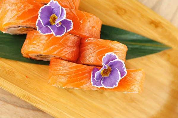 Suşi ruloları canlı somon dilimleri ve ahşap bir tabakta ustalıkla dizilmiş yenilebilir çiçekler içerir. Yemek Japon yemek sunumunu ve becerisini vurguluyor.
