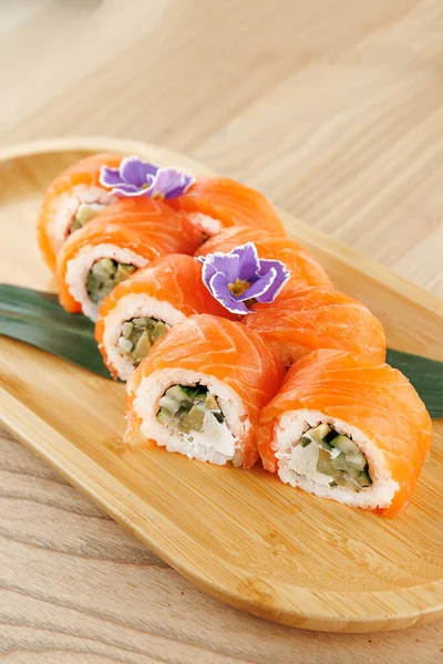 Somon suşi ruloları ahşap bir tabakta güzelce düzenlenir. Her rulo sebze ve pirinç içeriyor, üstünde süs çiçekleri var. Ayarları doğal ve basit..