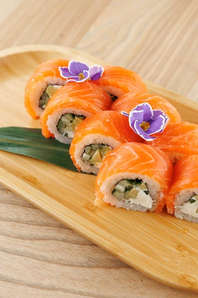Altı suşi rulosu ahşap bir tabakta sergilenir. Her rulo somona sarılmış ve salatalık ve balıkla dolu. Taze çiçekler sunuma renk ekler.