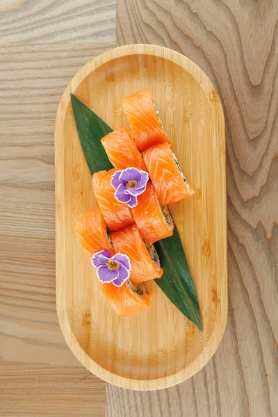 Süslü yeşil yapraklar ve mor çiçeklerle ahşap tepside dizilmiş somon balıklı suşi dürümleri. Bu yemek Japon mutfağını çağdaş bir şekilde sergiliyor..