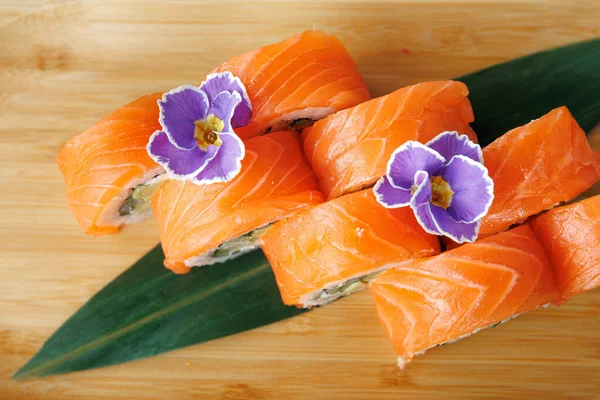 Suşi ruloları bambu bir tahtada güzelce düzenlenir. Her rulo somon içerir ve mor çiçeklerle süslenir. Yemek masası ya da sıradan bir yemek için mükemmel..