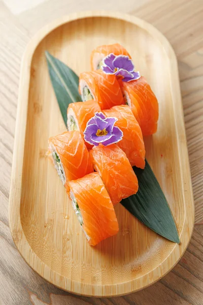 Somondan yapılmış taze suşi ruloları, ahşap bir tabağa konur. Tabakta yeşil yapraklar ve dekorasyon için canlı yenilebilir çiçekler yer alıyor. Yemek veya atıştırmalık için mükemmel..