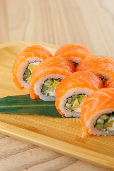 Ahşap bir tabakta avokadoyla doldurulmuş ve taze somona sarılmış suşi ruloları sergileniyor. Bu yemek, canlı renkler ve temiz sunumlarla geleneksel Japon mutfağını vurgular..