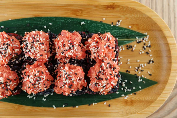 Suşi börekleri, üzerine baharatlı somon karışımı ve susam tohumları serpiştirilmiş ahşap bir tabakta oturur. Tabakta garnitür için yeşil yapraklar var. Renkli bir yemek sunuyor..