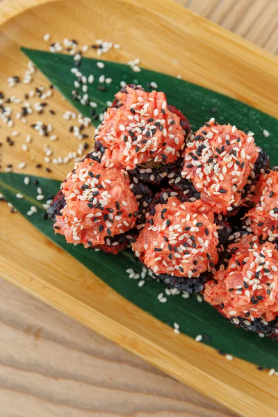 Farklı suşi çeşitleri yeşil yapraklar üzerinde sıralanır, beyaz ve siyah susam tohumlarıyla süslenmiş parlak pembe ve siyah pirinç topları sergilenir. Bambu tabağı doğal bir dokunuş katıyor..