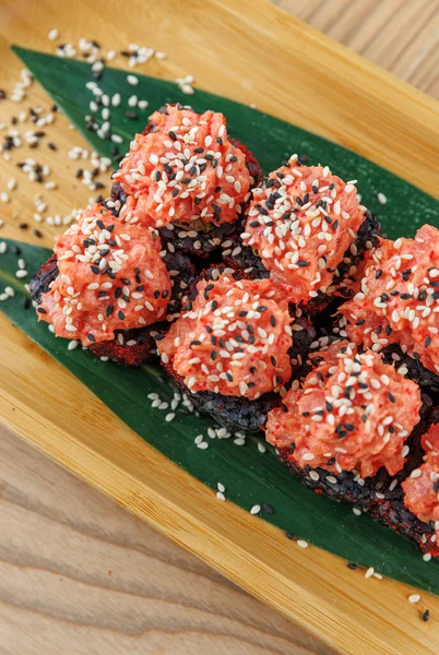 Canlı malzemelerle yapılan suşi ruloları ahşap bir tahtanın üzerinde sergilenir. Ekmeğin üzerinde pembe bir sos var ve susam tohumları serpiştirilmiş. Yeşil bir yaprak renk katar.