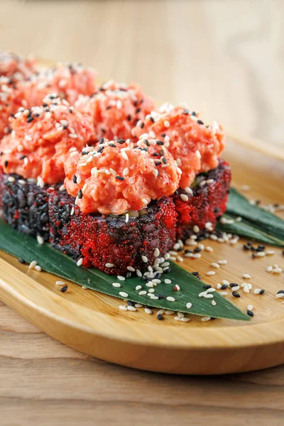 Suşi ruloları ahşap bir tabakta güzelce düzenlenir. Ekmekler somon ve susam tohumlarıyla kaplı. Dekoratif yeşil yapraklar sunuma biraz renk katar.