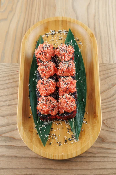 Suşi ruloları üzerine susam tohumları serpiştirilmiş bambu bir tabakta düzenlenir. Çanak, ahşap masa yüzeyinde renkli bir sunum sergiliyor..