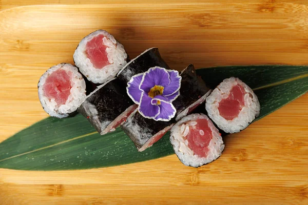 Bir bambu tabağında ton balığıyla dolu beş suşi rulosu düzenlenir. Mor bir çiçek, Japon mutfağında kullanılan taze malzemelerin altını çizerek yemeğe renk katar..