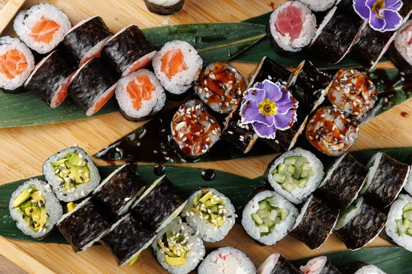 Çeşitli suşi ruloları ahşap bir tabakta düzenlenir, farklı balık ve sebze dolguları sergilenir. Yemek garnitür ve sosları içeriyor, çekici bir sunum hazırlıyor..