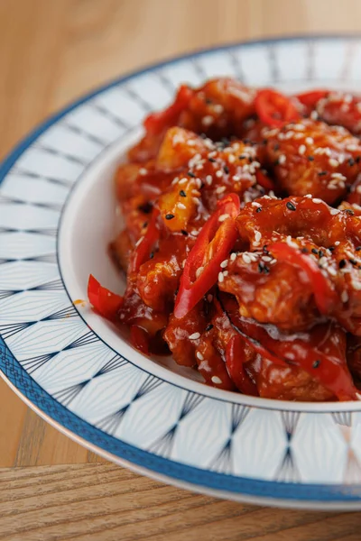 Bir tabakta dilimlenmiş kırmızı biber ve susam tohumlarıyla baharatlı kırmızı bir sosta pişirilmiş karides bulunur. Tabak, ahşap masalı canlı bir restoranda servis ediliyor..