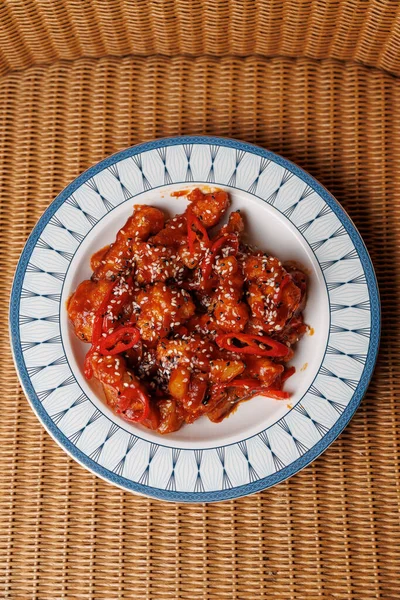 Susam tohumu ve kırmızı biberle süslenmiş baharatlı sosta pişirilmiş bir gurme tavuk tabağı dekoratif bir tabakta sunulur. Ayarda hasır kaplamalı bir arkaplan var.