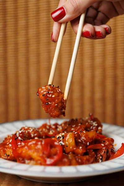 A hand holds chopsticks picking up a piece of fried chicken coated in sweet and sour sauce and served with colorful bell peppers and sesame seeds on a plate.