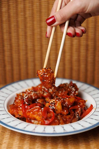 Hand holds chopsticks to pick up a piece of chicken topped with sauce and sesame seeds from a bowl filled with mixed vegetables.
