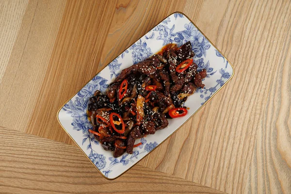 Bir tabakta dilimlenmiş kırmızı biber ve susam tohumlarıyla karıştırılmış kızarmış sığır eti var. Tabakta ahşap bir masada dekoratif bir tabakta servis edilen tatların bir karışımı sergileniyor..