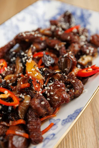 Dilimlenmiş sığır eti canlı biber, mantar ve süslemeli mavi ve beyaz bir tabakta susam tohumlarıyla kızartılır. Tabakta zengin lezzetler ve dokular sergileniyor.