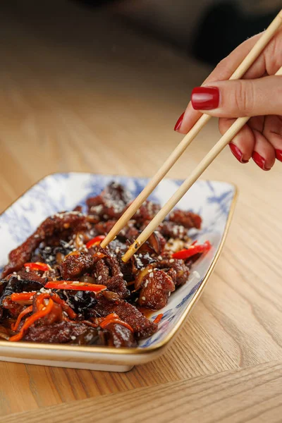 A hand holds chopsticks to pick up pieces of meat and vegetables from a beautifully arranged dish on a wooden table. The focus is on the vibrant colors and textures of the food.