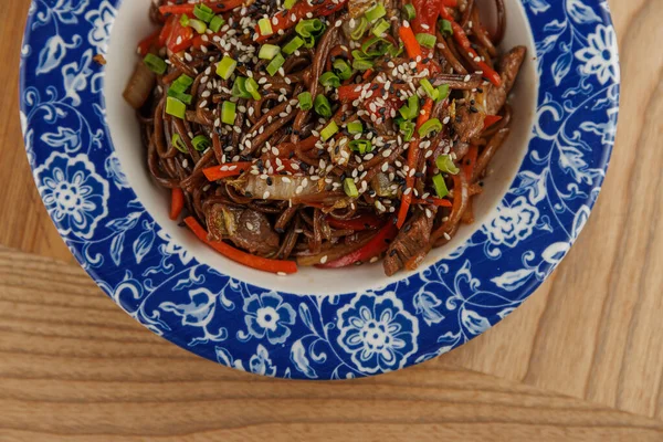 Bir kase kızarmış şehriye renkli sebzeler ve dekoratif bir tabakta sıralanmış susam tohumları içerir. Bu yemek Asya mutfağından bir tat sunuyor..