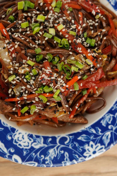 Kırmızı biber ve soğan gibi renkli sebzelerle karıştırılmış soba erişteleri, susam tohumlarıyla kaplı bir tabakta bulunur. Yemek canlı ve davetkar görünüyor..