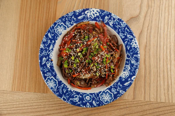 Erişte ve sebzeler mavi bir kasede birlikte kızartılır. Tabak susam tohumları ve otlarla süslenmiş, tahta bir masa yüzeyinde dinleniyor..