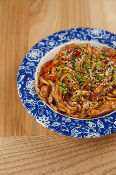 Bir kasede rengarenk sebzelerle karıştırılmış ve susam tohumuyla kaplanmış ramen var. Tabak parlak bir ortamda ahşap bir masanın üzerinde duruyor..