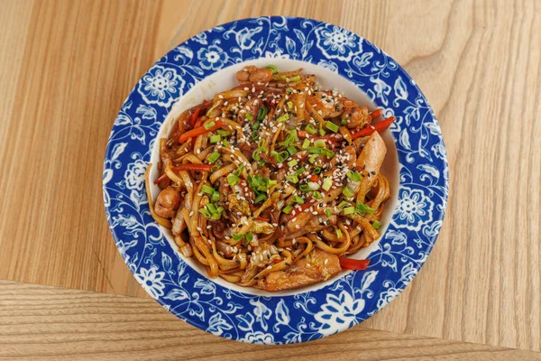 Bir tabakta bol miktarda kızarmış şehriye, tavuk ve canlı sebzelerle karıştırılmış, susam tohumları ve otlarla kaplı, ahşap bir masaya konmuş..