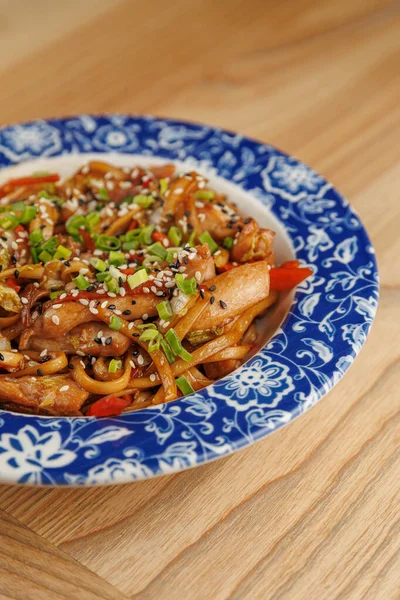 Erişteler yeşil soğan, susam tohumu ve kırmızı biberle kaplı mavi desenli bir kaseye konur. Tabak ahşap bir masada sergileniyor..