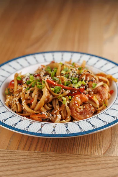 Bu sahnede karides, renkli sebzeler ve dekoratif bir tabakta servis edilen susam tohumlarının yer aldığı bir tabak erişte kızartması sergileniyor. Füzyon yemeğinin tadını çıkarın..