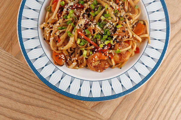 Bir kasede rengarenk sebzelerle karıştırılmış ve susam tohumuyla kaplanmış ramen var. Tabak, ahşap bir masadaki dekoratif bir tabağa konuyor ve canlı renklerini sergiliyor..
