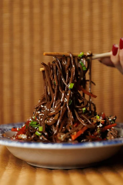 Erişte ve renkli sebzeler bir kasede karıştırılır. Çubuklar erişteyi kavrıyor, yemeye hazır. Bu yemek Asya mutfağını temsil ediyor..